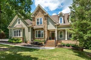 The large, two-story suburban house at 521 Belmont Circle in Wake Forest boasts green siding with stone accents and is surrounded by lush greenery and a manicured lawn. Multiple windows adorn the home, with a central staircase leading to the grand entrance.