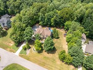 Aerial view of a suburban neighborhood with a large house surrounded by lush green trees. The house features a spacious yard with a driveway and a pool in the back. Other homes are visible nearby, integrated into the wooded landscape.
