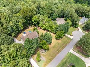 Aerial view of a suburban neighborhood surrounded by dense trees. A house with a backyard pool is visible in the foreground, situated at the end of a cul-de-sac. Neatly manicured lawns and driveways can also be seen.