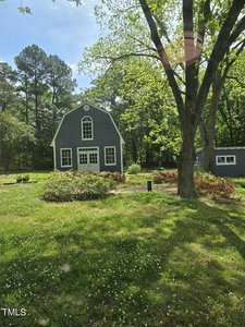 A charming, dark gray house with white trim stands amid a lush green yard filled with trees and bushes. The two-story structure features a large arched window and is surrounded by a peaceful, wooded landscape under a clear blue sky.