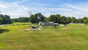 A spacious open field with a small house surrounded by trees. The house has a solar panel on the roof and several structures nearby. Power lines and a clear blue sky are visible in the background.