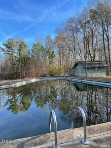 A quiet outdoor swimming pool with metal ladder rails leads into the water, reflecting the bare trees and blue sky of Oxford. To the right stands a small green shed at 115 Robin Road. The area is fenced and surrounded by trees, offering a peaceful retreat from city life.
