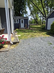 A gravel path leads to a small gray building surrounded by trees. The building has a window and a wooden deck with steps. On the left, part of a larger building is visible, and to the right, there's a gray garage. Bright green grass and bushes adorn the scene.