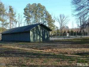 A small, green wooden shed stands in an open grassy area. Behind it, a tennis court is visible, enclosed by a chain-link fence. Tall trees and clear blue sky form the backdrop.