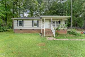 A single-story house with light siding and dark shutters, featuring a small front porch with steps. It's surrounded by a green lawn and trees in the background. A red lawnmower is visible to the right near the edge of the yard.
