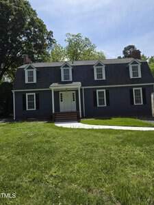 A two-story, dark gray house at 511 Providence Road, Oxford, boasts a steep shingled roof and white-trimmed windows. The front features a central entrance with a small porch and a pathway cutting through the lush green lawn, all embraced by trees under a clear sky.