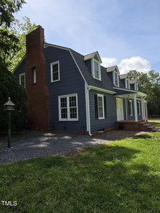 A blue house with dormer windows and a red brick chimney stands in a grassy area. A gravel path leads to the porch, and trees surround the house under a partly cloudy sky.