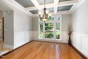 A bright dining room with a coffered ceiling and chandelier. The room has wainscoting, hardwood floors, and large windows offering a view of a garden. A decorative vase with branches stands in the corner.