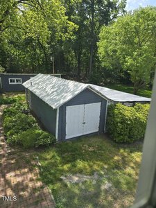 A small blue-gray shed with white doors is surrounded by lush greenery and trees. A brick walkway leads to and past it, while another building is visible in the background. The scene is lit by bright sunshine.