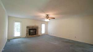 A spacious, empty living room with light beige walls and a textured ceiling. It features a ceiling fan with lights and a stone fireplace. Two tall windows on the left and right allow natural light into the room. The carpet is light blue.
