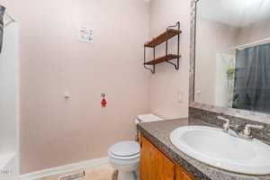 A small bathroom with a granite countertop, white sink, and wooden cabinets. There's a toilet next to an empty wall space for storage. Above, a set of shelves is mounted. A shower is partially visible with a curtain partially open.