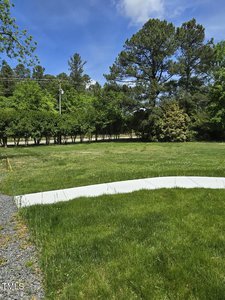 A lush, green lawn with a white curved pathway leads to a row of trees under a partly cloudy blue sky.