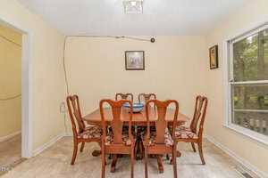 A dining room with a wooden table and six matching chairs, each with floral-patterned cushions. A framed picture hangs on the wall above the table, and a window to the right shows trees outside. A small dish is centered on the table.