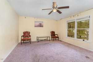 A sparsely furnished living room with two armchairs, a glass coffee table, a ceiling fan, and a large window letting in natural light. The walls are painted light, and the carpeted floor is beige. A wall decoration hangs between the chairs.