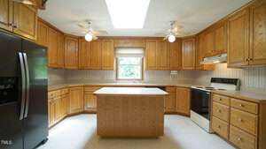 A spacious kitchen with wooden cabinets, a central island, and a white countertop. It features a black refrigerator on the left, and a white stove and oven on the right. Two ceiling fans and a skylight provide lighting. A window is above the sink.