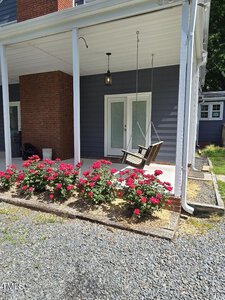 A porch swing hangs from a covered patio near a brick wall. Bright red flowers are planted in a neat line along the edge of the patio, bordered by gravel. Double glass doors and an exterior lamp are visible in the background.