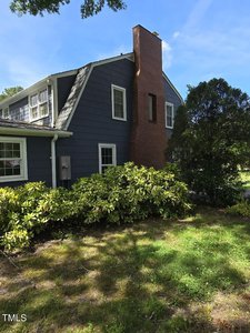 A two-story house with dark gray siding and a red brick chimney is surrounded by green shrubs and trees. The sky is clear and blue, casting shadows on the grass in the yard.