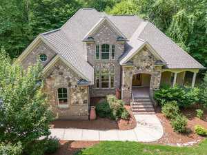 A large, two-story brick and stone house with a gabled roof is surrounded by lush greenery and trees. A curved walkway leads to the front entrance, flanked by shrubs and potted plants. The house has multiple windows and detailed stonework.