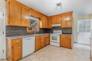 A kitchen with wooden cabinets, a window above a double sink, a stove with an overhead fan, and a dishwasher. The floor is light-colored, and there's a back door to the right. The walls are partially tiled with a patterned backsplash.