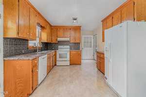 A spacious kitchen with wooden cabinets, a white refrigerator, dishwasher, and stove. The floor is light-colored, and a door leads to another room. A window provides natural light, and a patterned backsplash adds detail above the countertops.