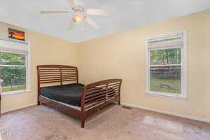 A sparsely furnished bedroom with beige walls featuring a large wooden bed with a black sheet. Two windows provide natural light, revealing a view of greenery outside. A ceiling fan hangs above, and a small piece of art adorns the wall.