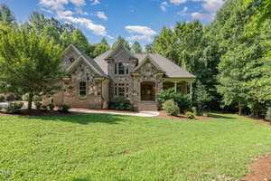 A two-story brick and stone house at 1180 Oak Grove Court, Wake Forest, boasts arched windows and is enveloped by lush greenery. The front entrance features a small porch with steps leading to a wooden door, set against the backdrop of a clear sky with a few clouds.