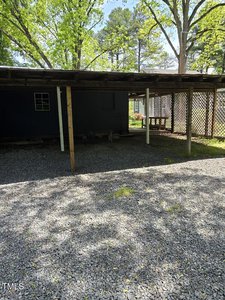 A shaded outdoor area with a wooden roof supported by posts, covering a section of gravel underneath. To the right, lattice panels partially enclose the space. Green trees and a building are visible in the background.