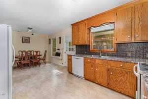 A kitchen with wooden cabinets and a tile backsplash on the right, featuring a dishwasher and oven. A dining area with a table and chairs is visible in the background. The room is bright with natural light coming through the window.