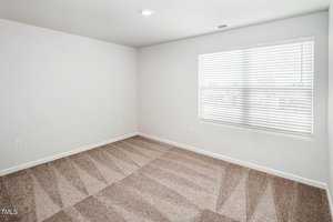 An empty room with light beige carpet and white walls. A window with closed blinds allows soft natural light to enter the space, creating a calm atmosphere. The room is unfurnished, providing a blank canvas for decoration.