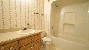 A bathroom featuring floral wallpaper, a wooden vanity with a white sink, and a toilet. The bathtub and shower are combined with built-in shelving. A towel bar is above the toilet.