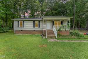 A small, single-story house at 215 E Quailridge Road features light siding and dark shutters, nestled amidst lush green grass and tall trees in Oxford. The charming home boasts a front porch with steps leading up to it, set against the serene backdrop of dense woods.