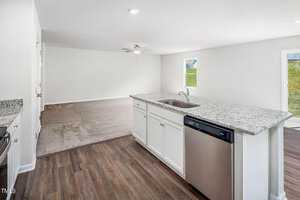 Modern open-concept kitchen and living area with light gray walls, a granite countertop island, and stainless steel appliances. The kitchen has wood flooring, while the adjacent living space is carpeted. A window and door provide natural light and access outside.