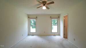 An empty room with white walls and carpet, featuring two windows with roller blinds. A ceiling fan with a light is centered above. A doorway leads to another room. Natural light filters in through the windows.