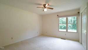 An empty room with light-colored walls and carpeting features a ceiling fan with a light. A large window with closed blinds overlooks a green outdoor space. A closed door is visible on the right wall.