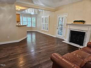A living room with wooden floors, a brown leather sofa, and a white fireplace. The adjacent open kitchen has wooden cabinets. Large windows with plantation shutters and a glass door allow natural light. Walls are painted beige.