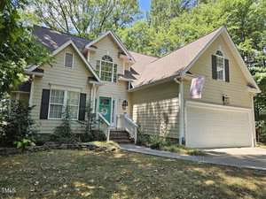 A charming two-story house at 563 Woodbridge Court in Wake Forest features light green siding, white trim, and a two-car garage. A small pathway leads to the front door, adorned with a decorative wreath. The home is enveloped by lush green trees and a well-kept lawn.