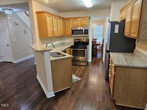 A modern kitchen with wooden cabinets, granite countertops, and stainless steel appliances, including an oven and refrigerator. The space features a double sink and hardwood flooring, with a view into a dining area through a doorway.