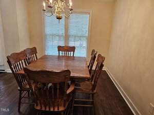 A wooden dining table with six matching chairs is set in a room with light yellow walls and dark hardwood floors. A decorative chandelier hangs above the table. Large white blinds cover the windows, partially letting in light.