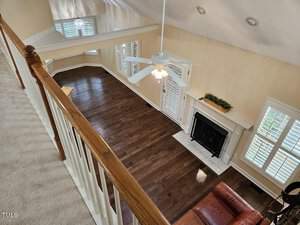 View from an upper floor overlooking a living room with dark hardwood floors, a white fireplace, and large windows with shutters. A ceiling fan hangs from the high ceiling, and a brown leather couch is partially visible.