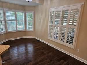 A room with hardwood floors features a large bay window with white plantation shutters, allowing natural light to enter. A window on an adjacent wall also has matching shutters. The walls are painted a light beige.