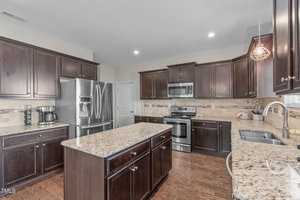 A modern kitchen with dark wooden cabinets, stainless steel appliances, and granite countertops. An island sits in the center. The room is well-lit with recessed lighting and features a hardwood floor.