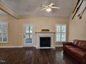 A cozy living room with a tan leather couch, a white fireplace, and wooden flooring. Two large windows with plantation shutters and a door provide natural light. Ceiling fan overhead, with decorative paddles and a wheel on the high walls.
