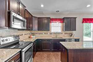 A modern kitchen with dark wooden cabinets, stainless steel appliances, and granite countertops. Red curtains adorn the windows, allowing natural light to illuminate the space. An island is centered in the room, and light-colored tiled backsplash is visible.