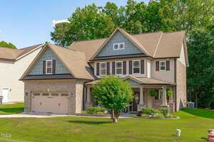 A two-story suburban house with a brick and siding exterior. It features a gabled roof, a double garage, and a covered front porch with hanging plants. The house is surrounded by a well-manicured lawn and bordered by trees in the background.