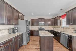 A modern kitchen with dark wood cabinets, granite countertops, and stainless steel appliances. A central island offers additional workspace. The flooring is wood, and a window with a red valance brings in natural light.
