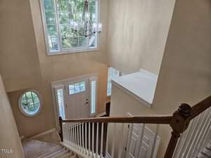 A staircase with white railings descends towards a front door with glass panels. Above, a large window lets in natural light, illuminating the beige walls and a hanging chandelier. A round window is visible on the left.