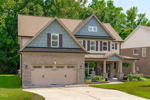 A two-story suburban house with a brick and blue siding facade, featuring a double garage, a small front porch with chairs, and a well-maintained lawn. The background includes dense green trees and a neighboring brick house.