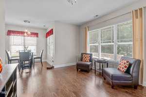 A bright dining and sitting area with wood flooring. The dining table has four chairs beneath a modern light fixture. Two cushioned chairs with colorful pillows and a small round table are near large windows with beige curtains.