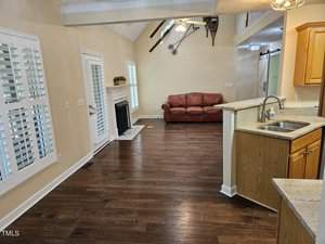 A living room with hardwood floors, vaulted ceilings, and a ceiling fan. It features a brown leather couch, a fireplace, and large windows with white shutters. The adjacent kitchen has a bar-style counter with wooden cabinets and granite countertops.