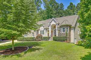 A suburban house with a light-colored exterior and arched entryway surrounded by green grass and trees. The landscaping includes trimmed hedges and a mulch bed with a small bush.